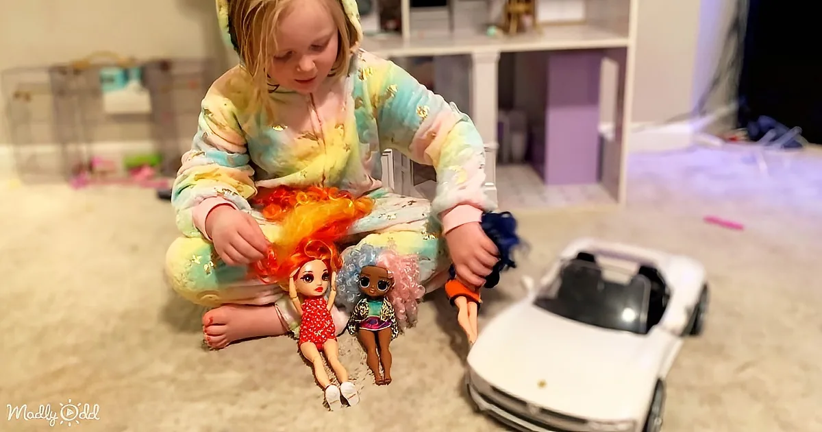 A child in tie-dye pajamas plays with dolls on a carpeted floor, with a toy car nearby.