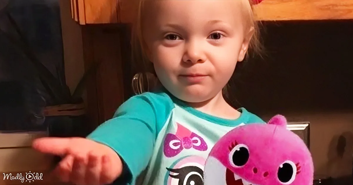 A young child wearing a turquoise shirt with a cartoon character holds a pink stuffed toy, extending their hand forward.
