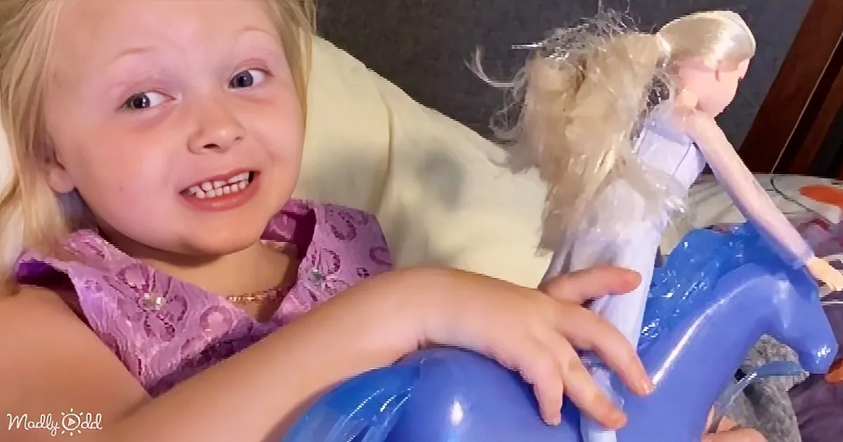 A young girl with blonde hair and a purple dress is holding a blue toy horse, smiling.