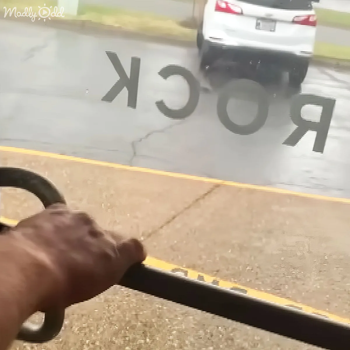 A hand grips a handlebar, with a blurry "ROCK" sign and a white car in the background.