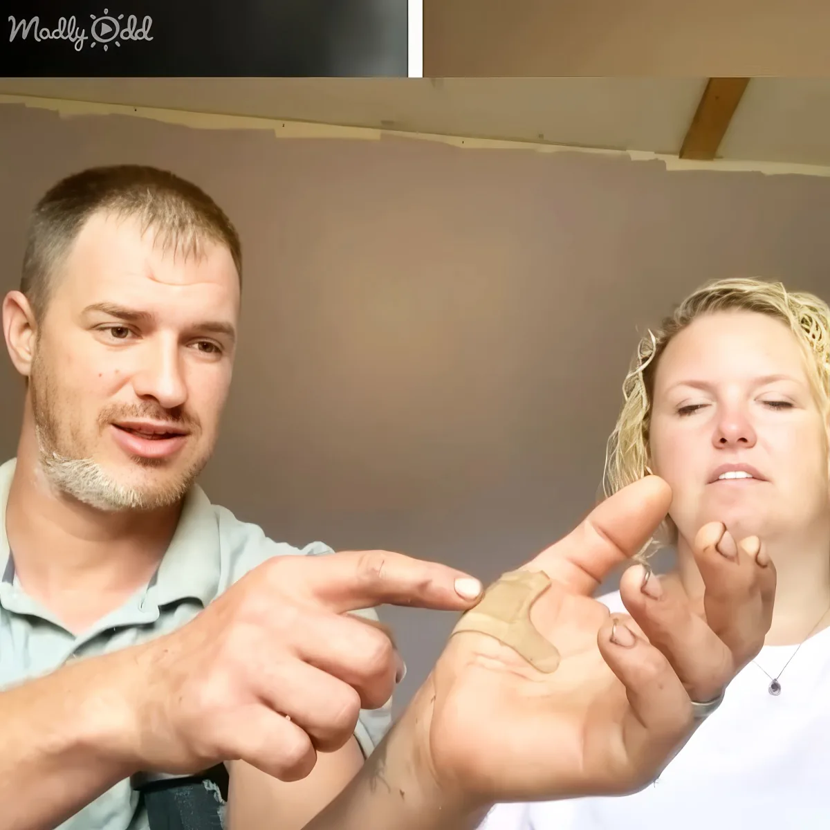 A man points at a woman's hand with a bandage, both appear to be in a room.