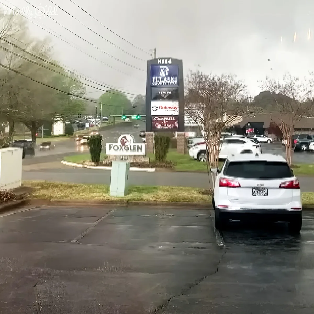 A white SUV is parked in a wet parking lot near a Foxglen sign and a large digital billboard.
