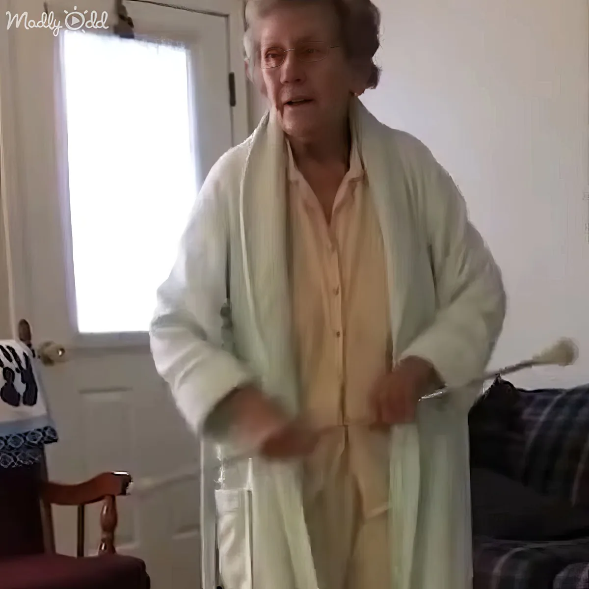 An elderly person in a white bathrobe stands indoors, holding a cane, with a door and window in the background.