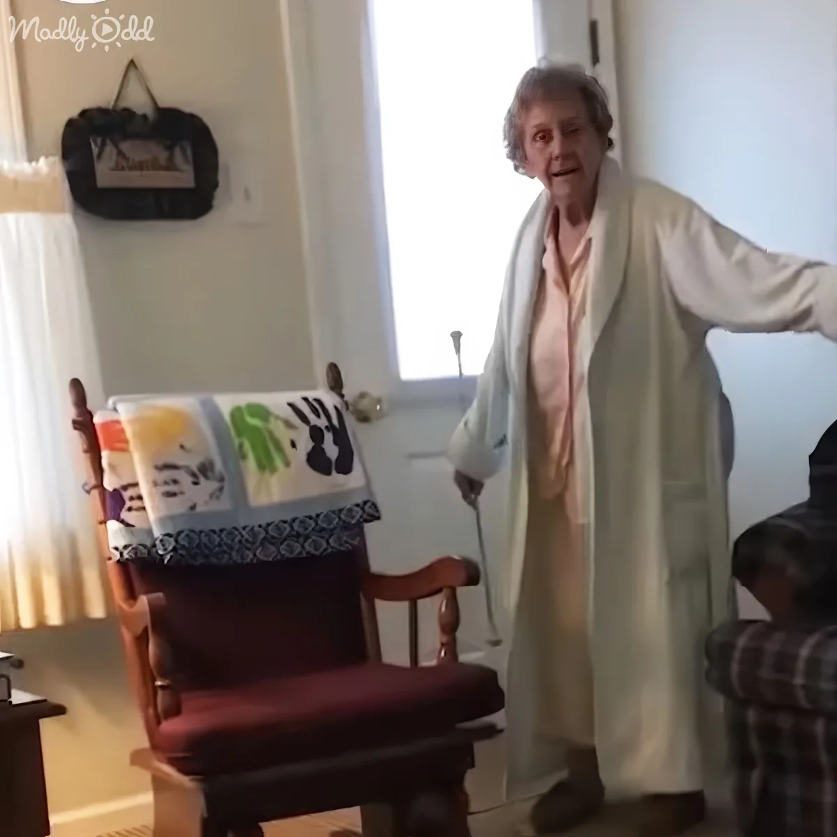 An elderly woman in a light robe stands in a room with a chair and a colorful towel draped over it.