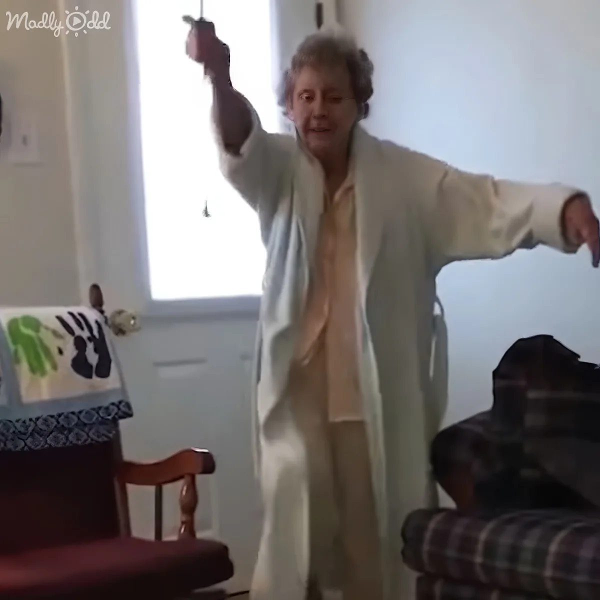 An elderly woman in a white robe is dancing joyfully in a room, holding a red object in her raised hand.