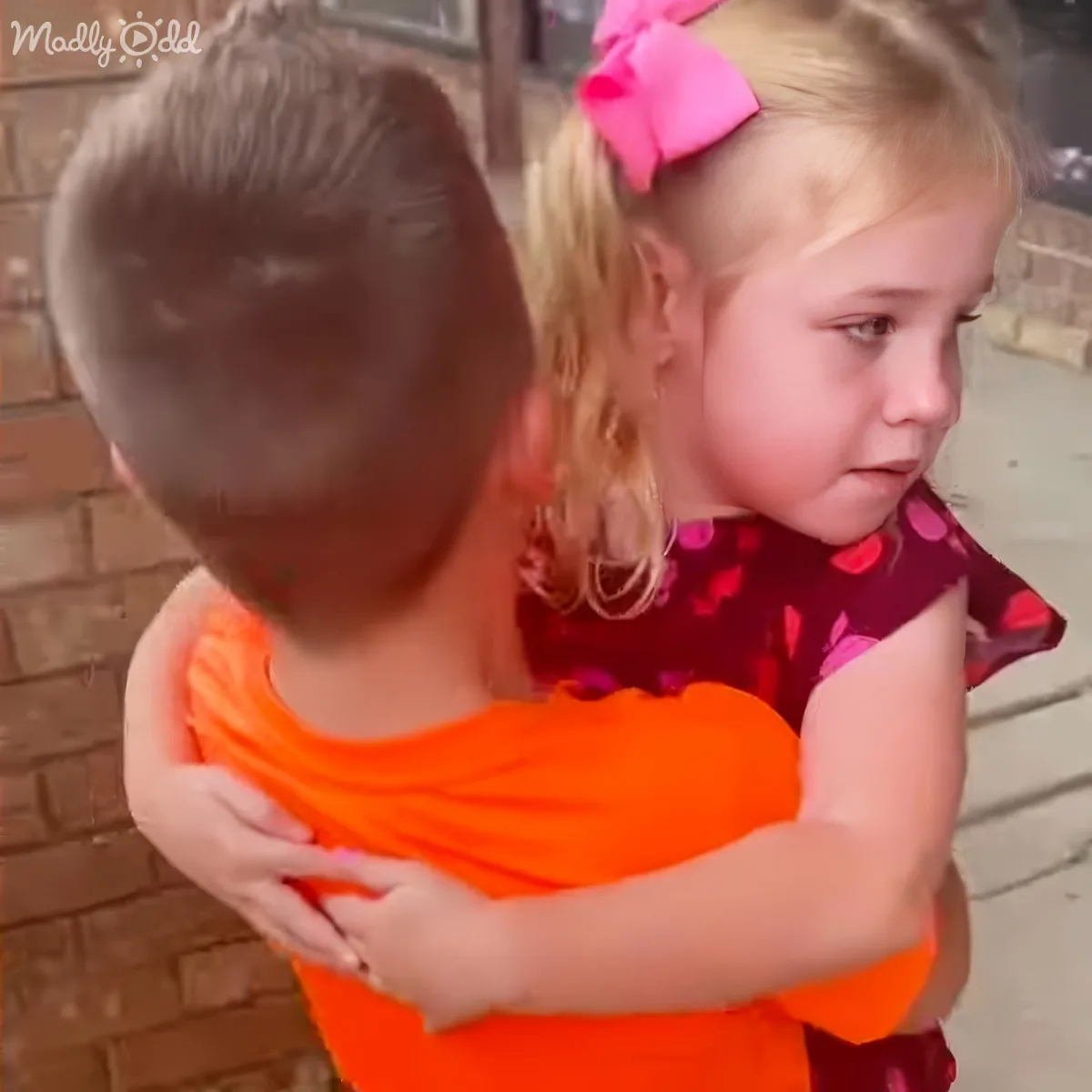 A child in an orange shirt is holding another child in a red dress with a pink bow.