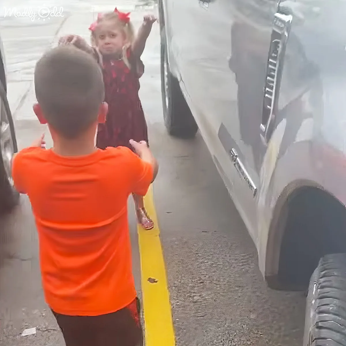 A young boy in an orange shirt holds a small girl in a red dress, both standing between parked cars.