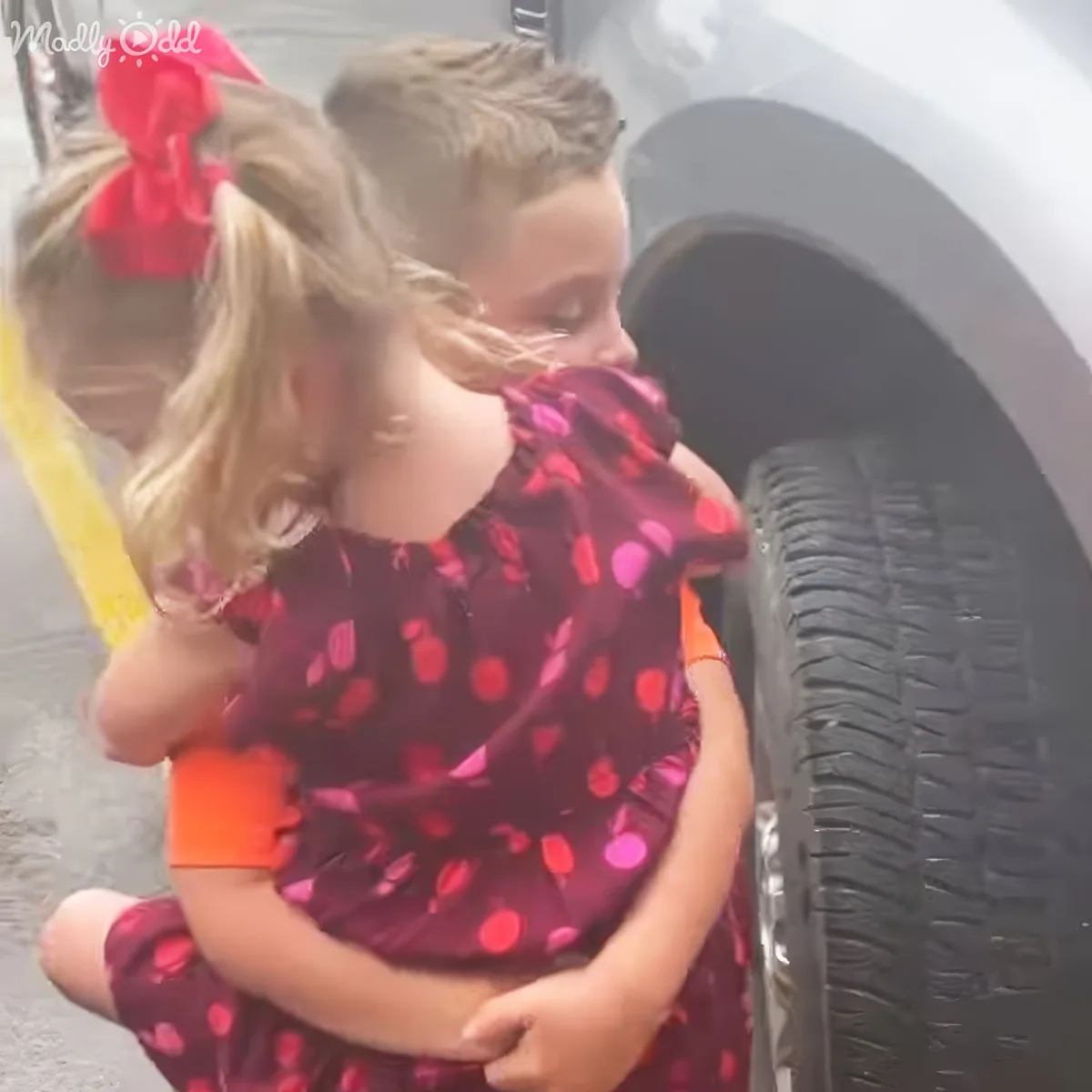 A young girl in a red polka-dot dress hugs another child near a car tire.