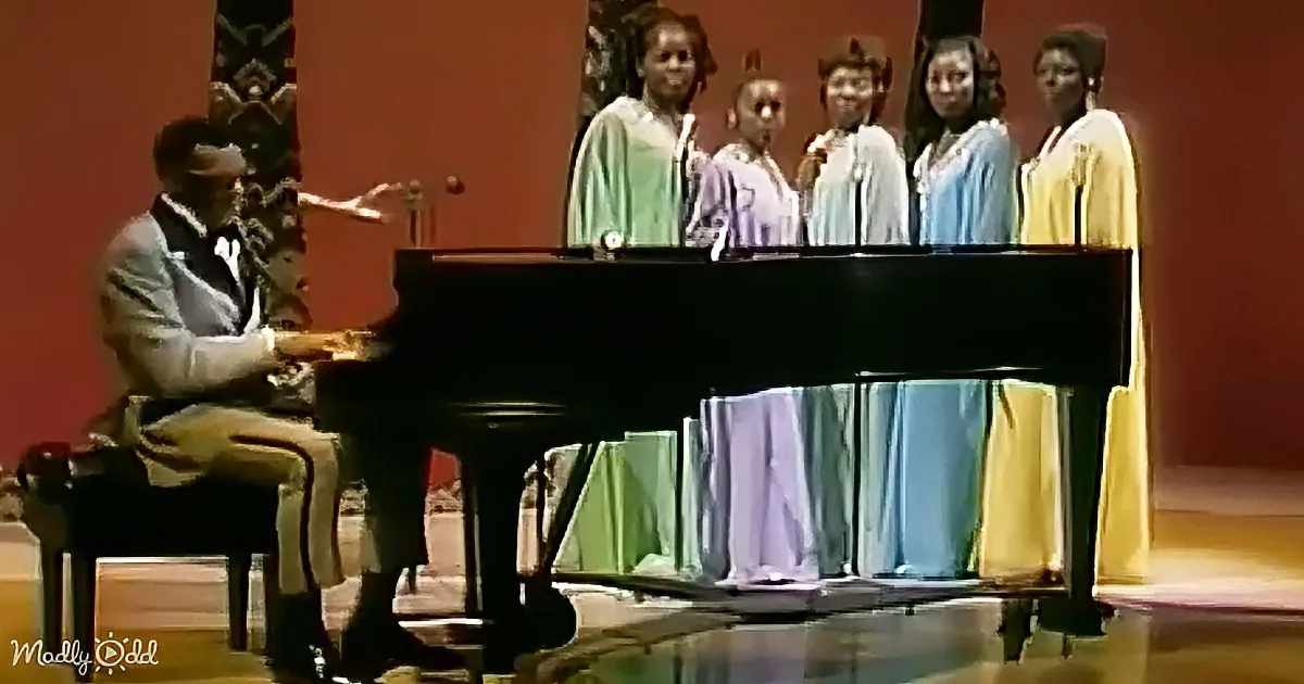 A man plays piano while a group of women in colorful robes sing behind him.