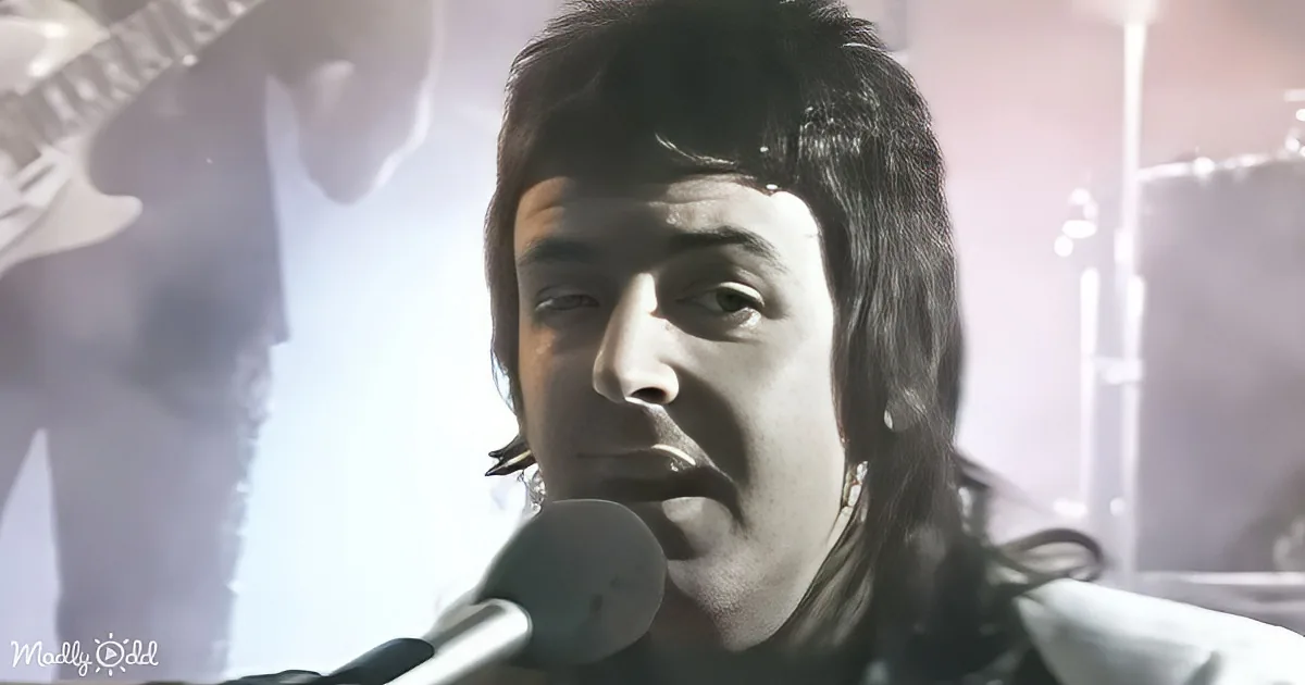 A person with long hair sings into a microphone, with a guitarist and drum set in the background.