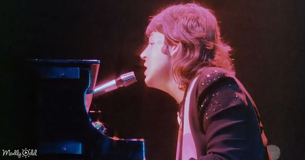 A person with long hair, wearing a sparkly shirt, is singing into a microphone while playing a piano.