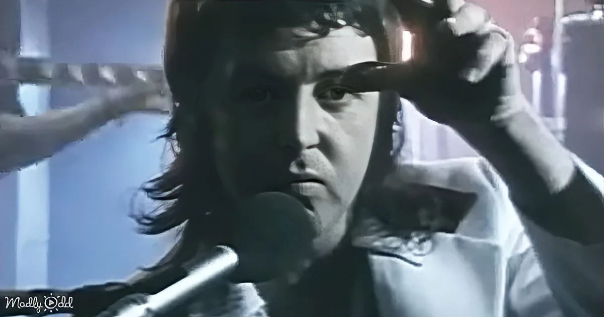 A person in a white shirt holds a microphone, adjusting their hair with one hand.