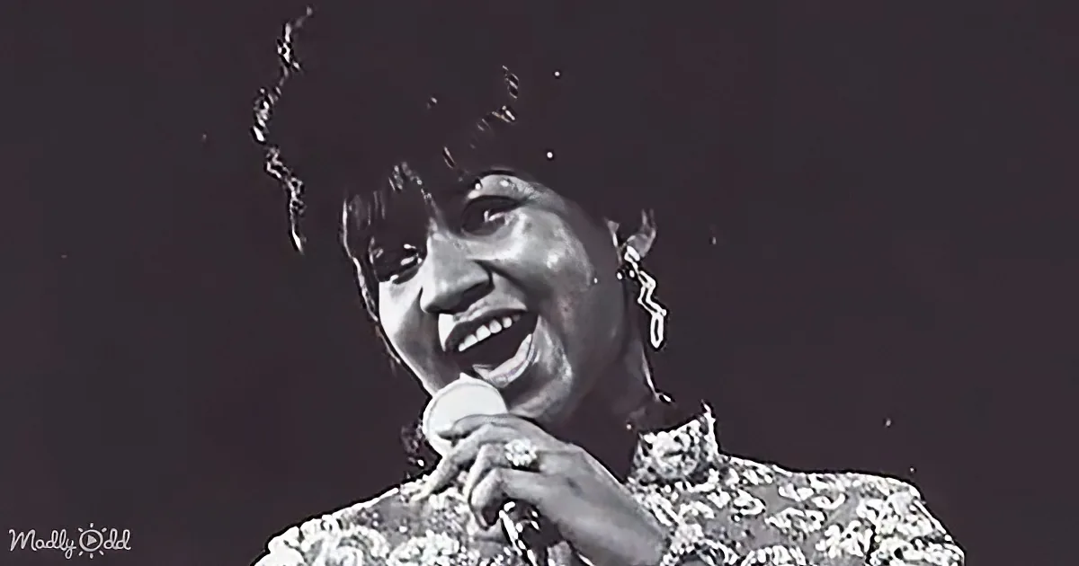 A woman is singing passionately into a microphone, wearing a patterned top and earrings, with a dark background.