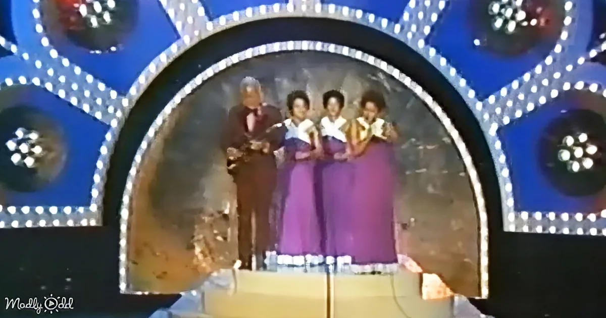 Four women in purple dresses stand on stage with a man in a suit, surrounded by bright lights and a blue backdrop.