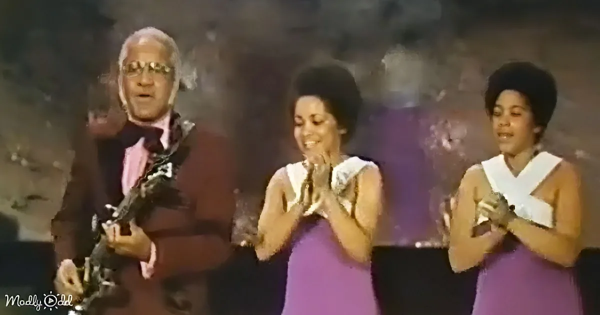 A man in a suit holds a trophy, two women in purple dresses clap, and smoke rises in the background.