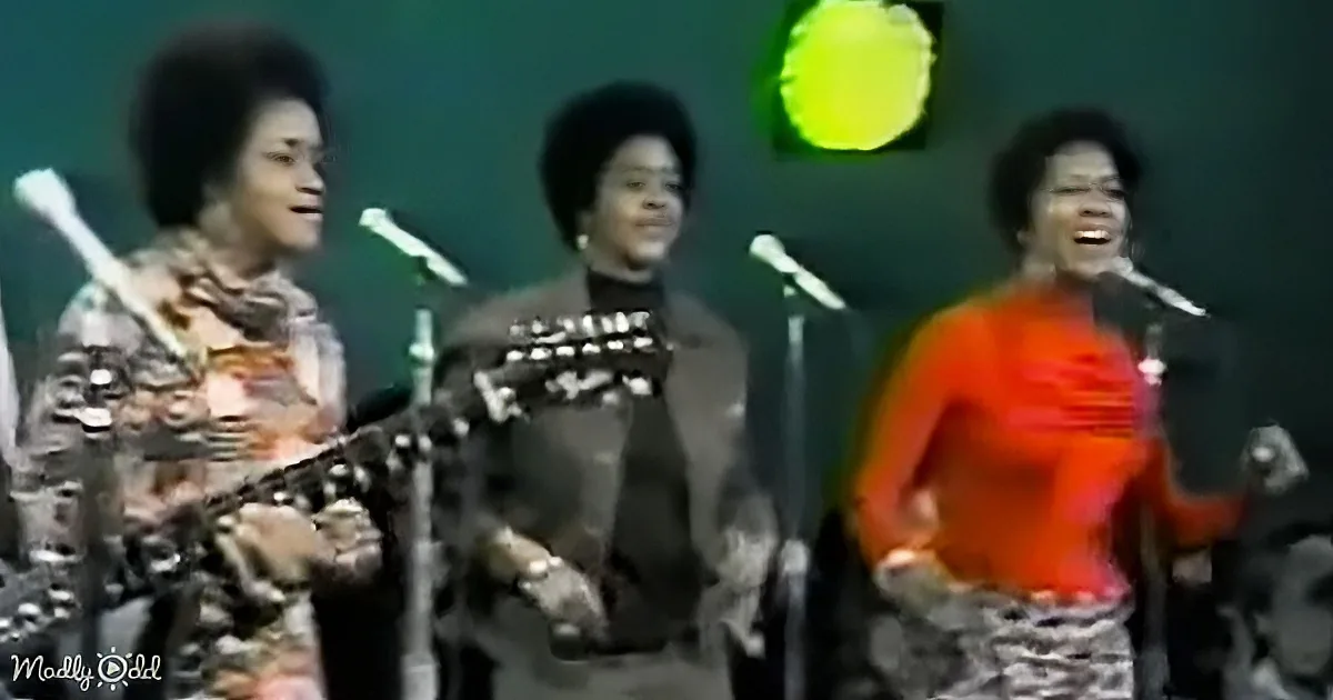 Three women singing on stage, one playing guitar, another with a microphone, and the third smiling.