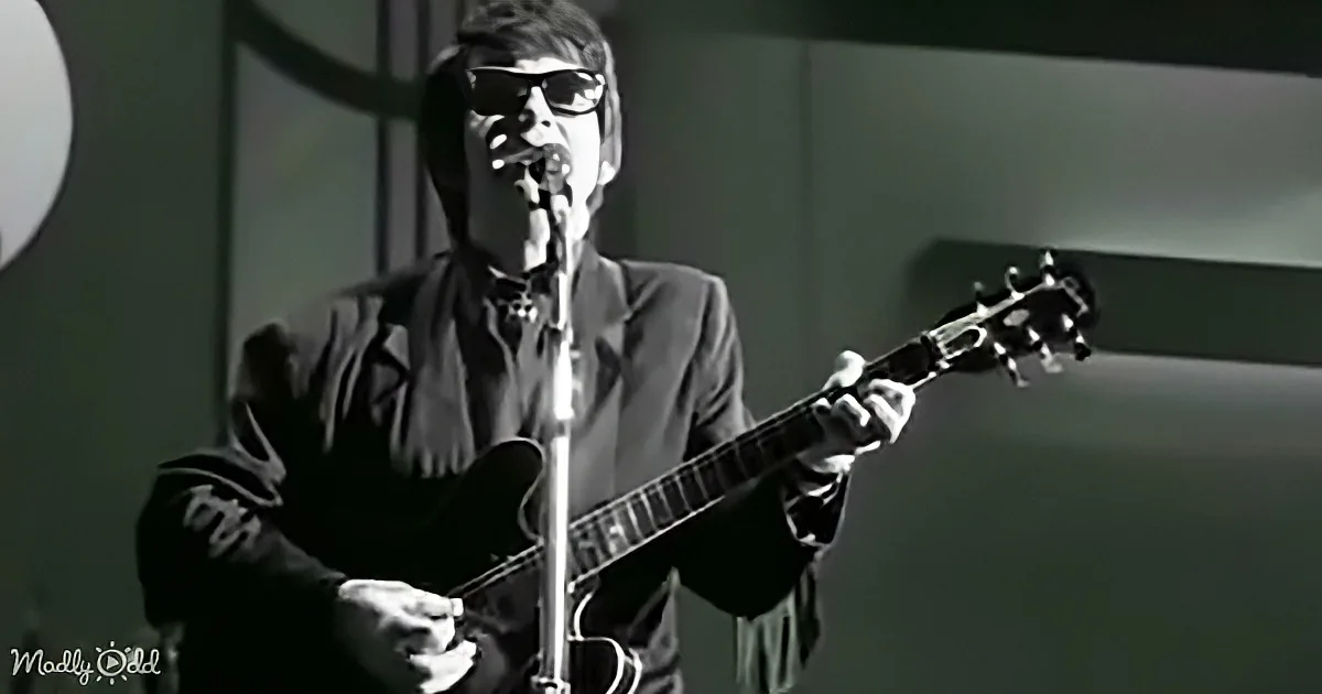 A musician in dark attire plays a guitar and sings into a microphone on stage.