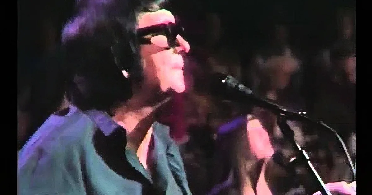 A person with short hair and sunglasses is holding a microphone, possibly singing or speaking.