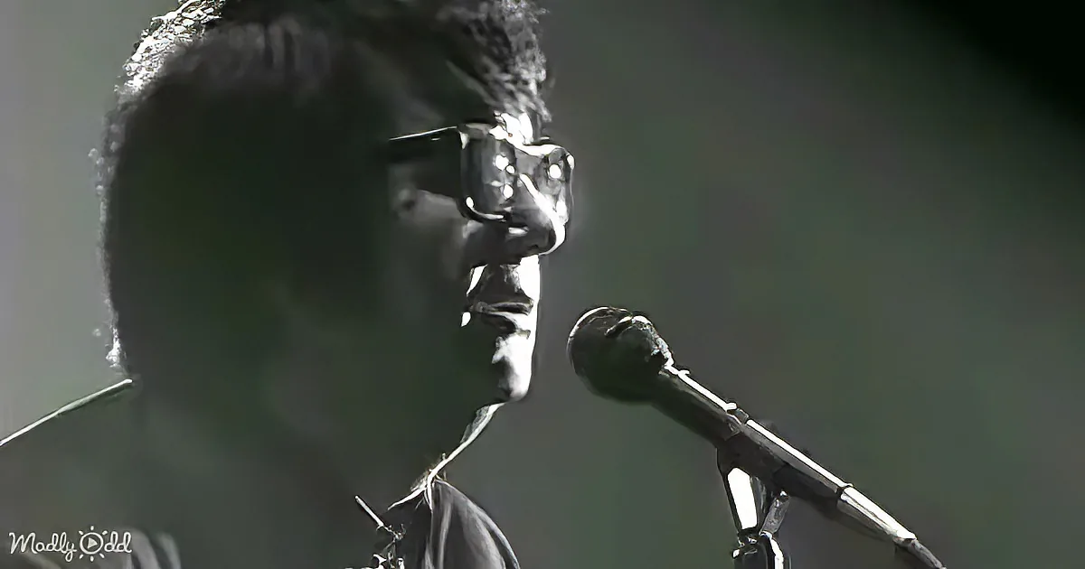 A person wearing glasses stands in front of a microphone, possibly singing or speaking.