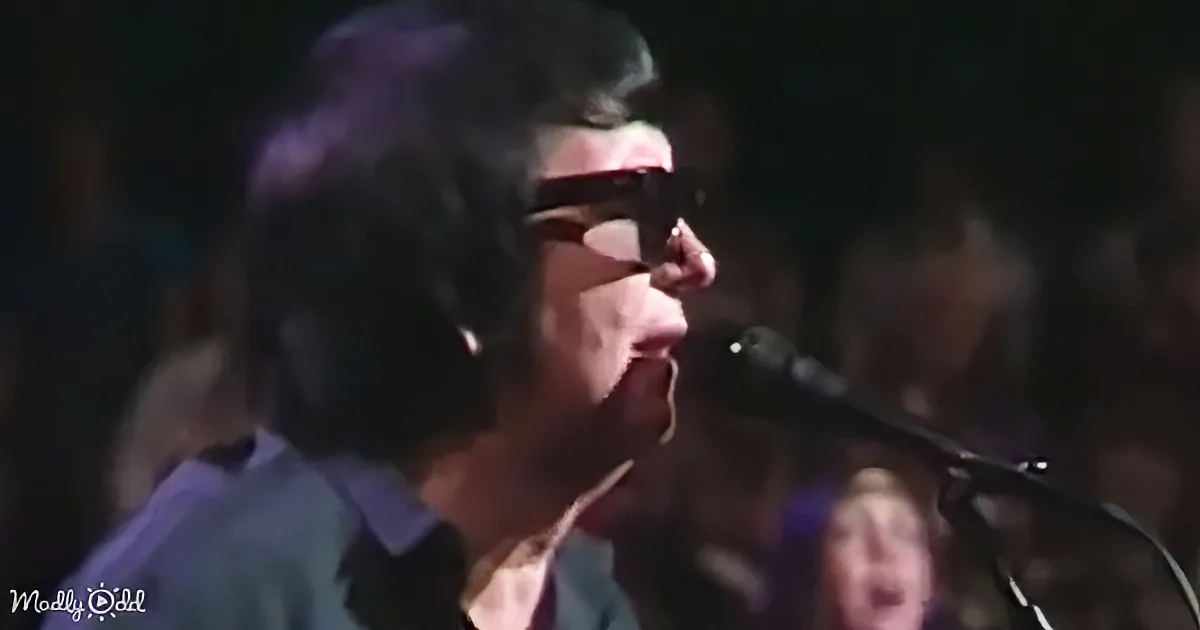 A person wearing sunglasses and a blue shirt sings into a microphone on stage.