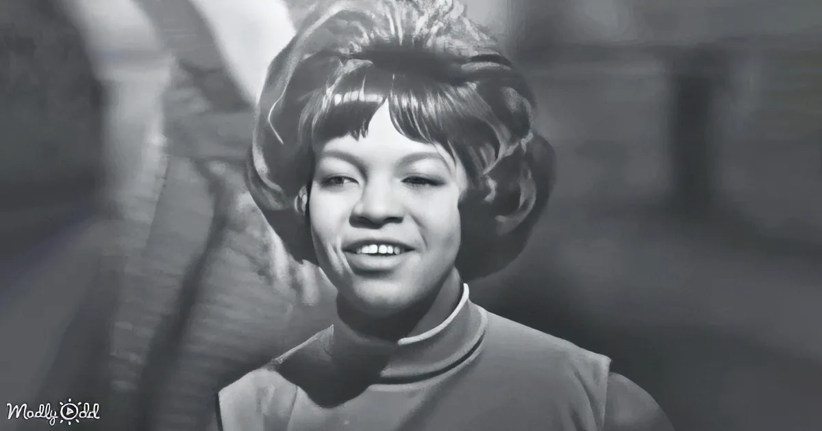 A smiling woman with short, styled hair and a high-collared outfit is the main subject of this black-and-white image.