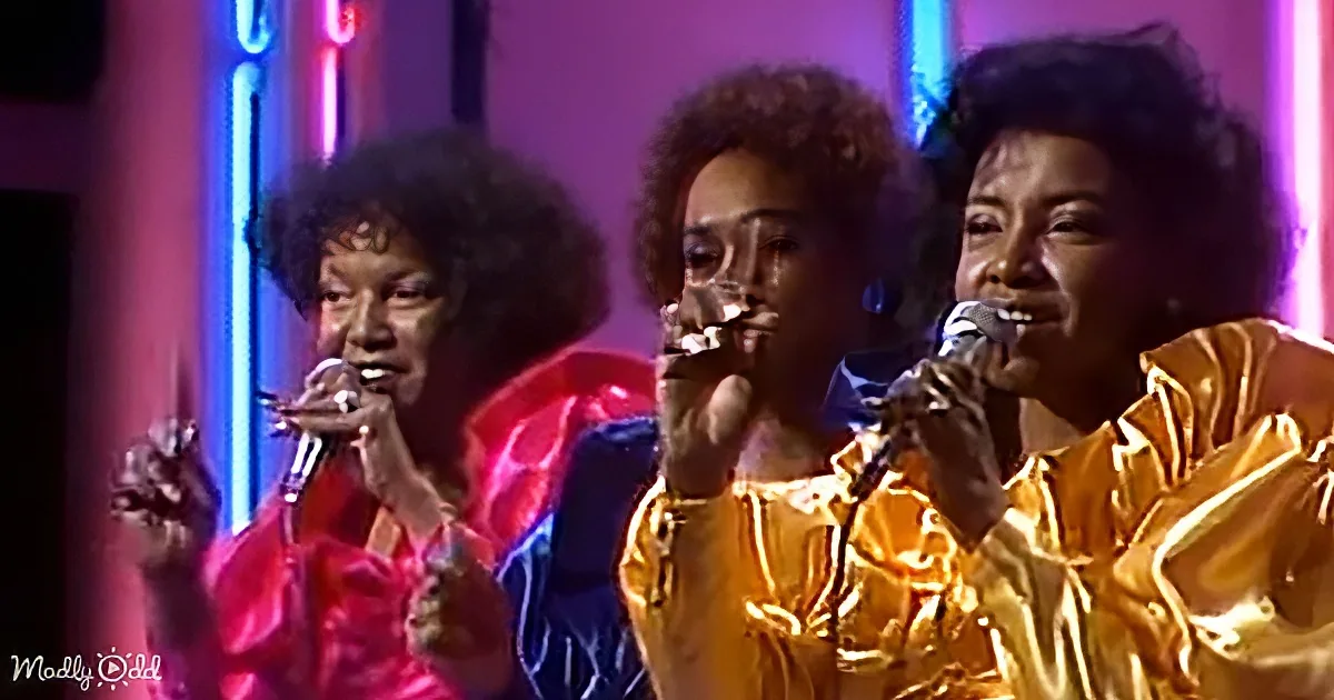 Three women in vibrant outfits sing into microphones on a stage with colorful lighting.