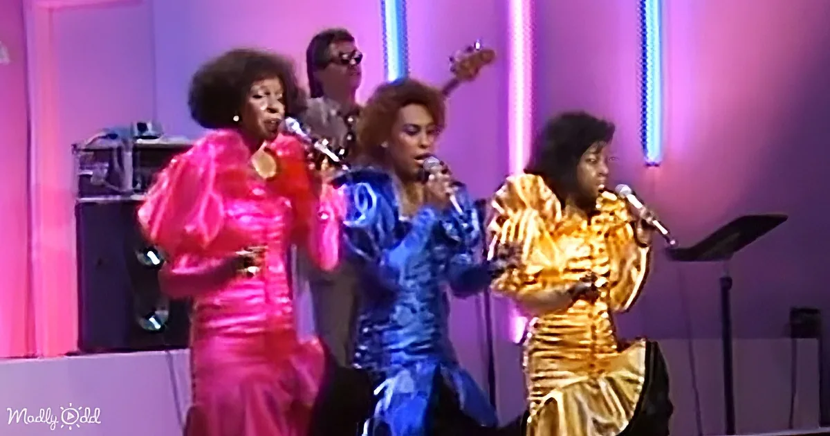 Three women in vibrant outfits sing on stage with a guitarist behind them.