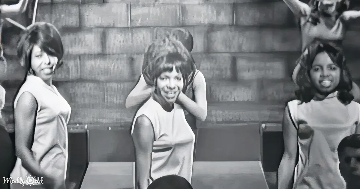 A woman in a sleeveless dress smiles while holding her hands behind her head, surrounded by other women in similar attire.