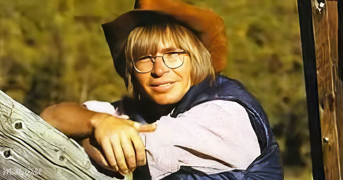 A person in a cowboy hat and glasses leans on a wooden fence, smiling.
