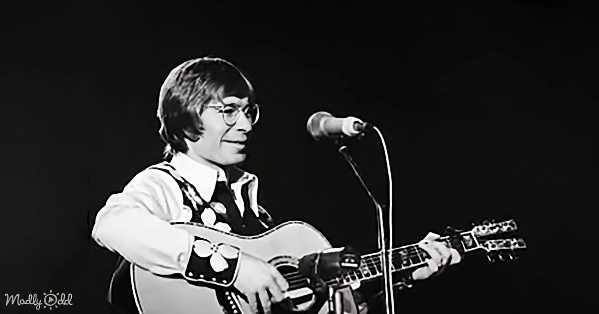 A person in a floral vest plays an acoustic guitar on stage with a microphone.