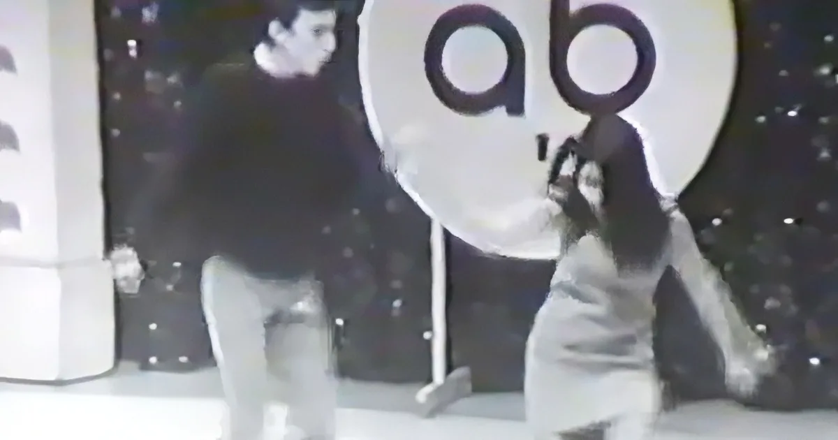 A person in a white outfit is dancing in front of a large "ab" sign with a starry background.