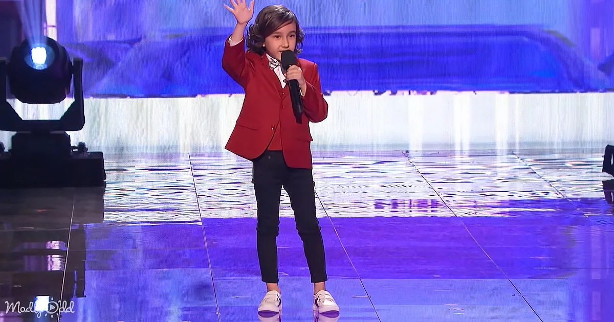 A child in a red blazer and black pants stands on stage, holding a microphone and raising one hand.