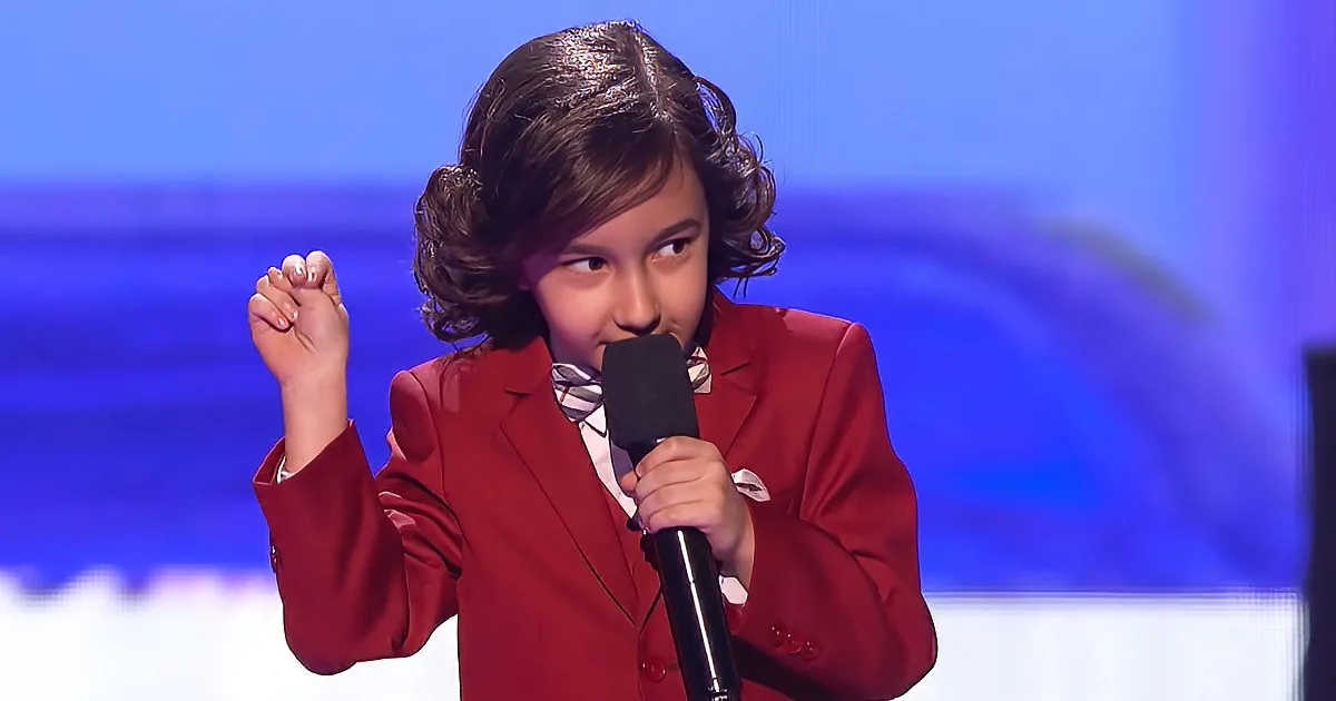 A young person in a red suit holds a microphone, looking to the side with a fist raised.