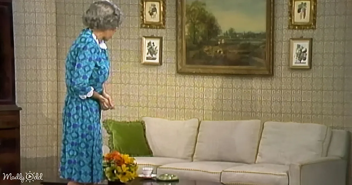 A woman in a blue dress stands near a white couch with green pillows, in a room with patterned wallpaper and framed pictures.
