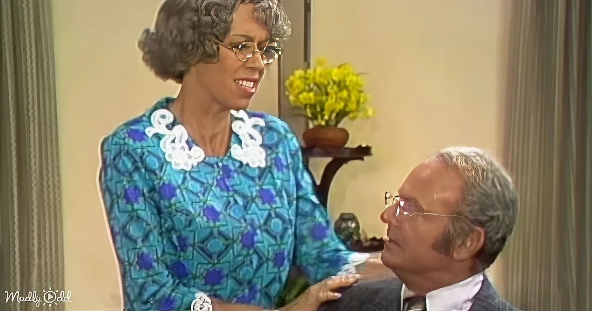 A woman in a blue patterned dress with white lace collar smiles at a man in a suit, both seated indoors.