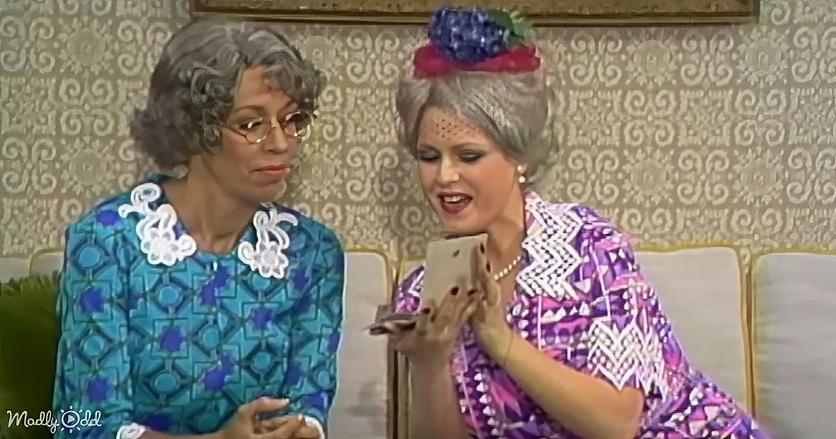 A woman in a colorful dress holds a small dog while another woman in a blue patterned dress looks on.