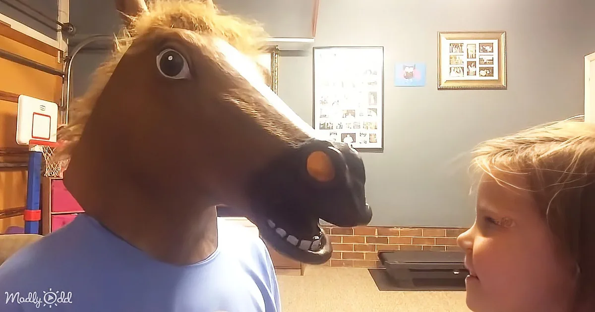 A person wearing a horse mask interacts with a child in a room with framed pictures and a basketball hoop.