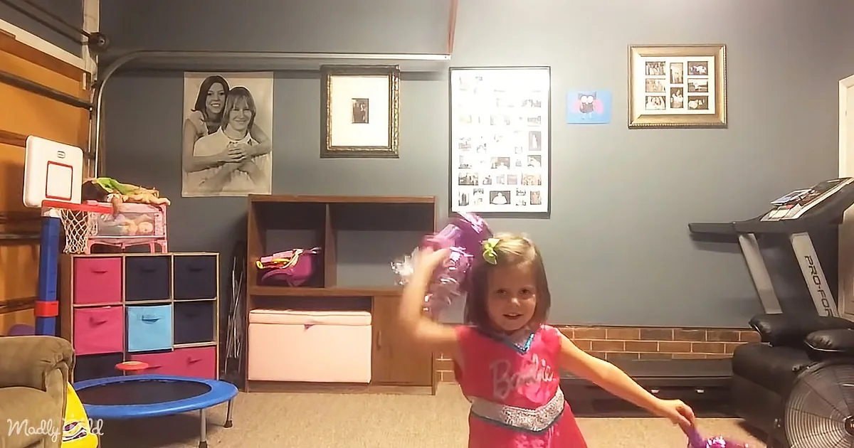 A young girl in a pink Barbie outfit dances in a room with toys, a treadmill, and family photos on the wall.