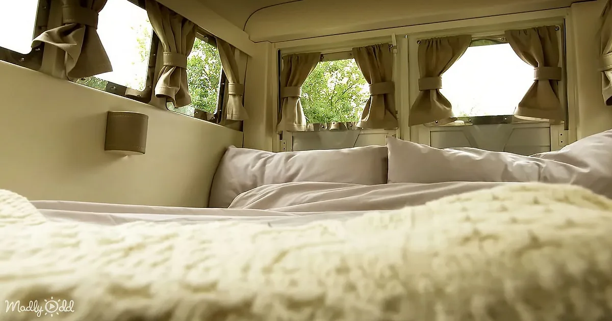 A cozy bed with white bedding and beige curtains in a van with multiple windows.