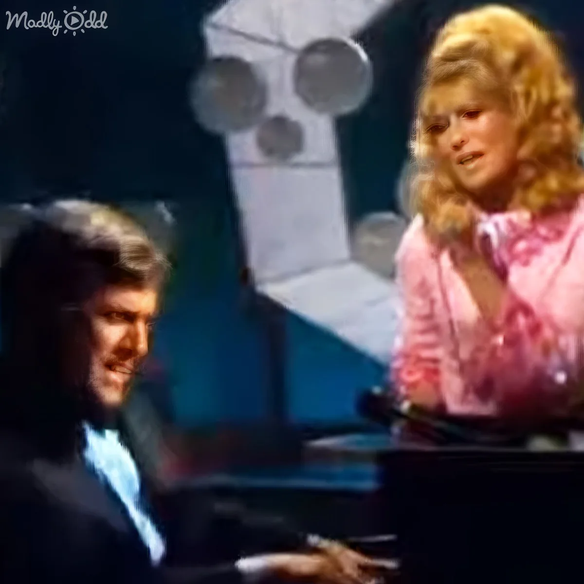 A man in a suit plays piano while a woman in a pink outfit sings beside him.