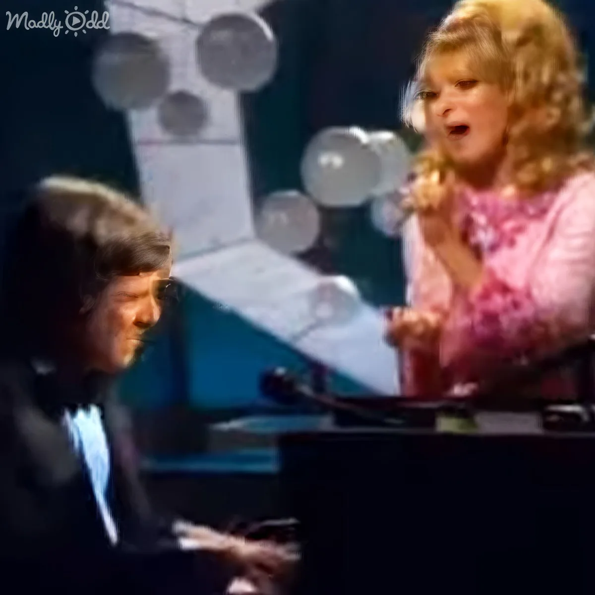 A woman sings passionately while a man plays piano, both on stage with microphones and sheet music.