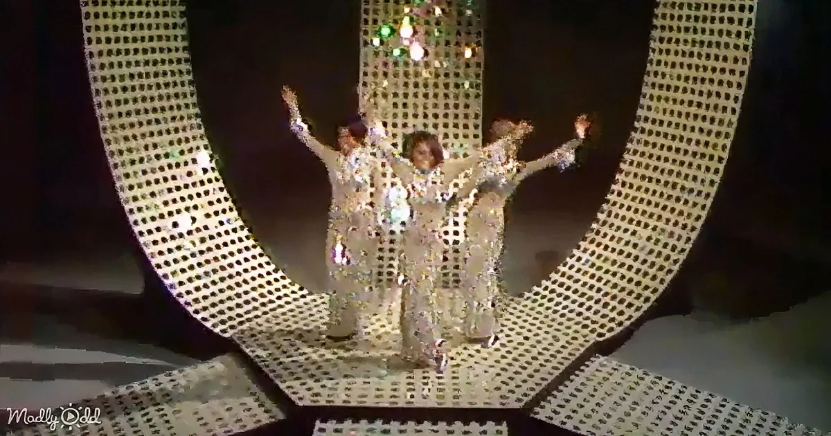 Three performers in sparkling silver outfits stand on a stage with a glittery backdrop, arms raised.