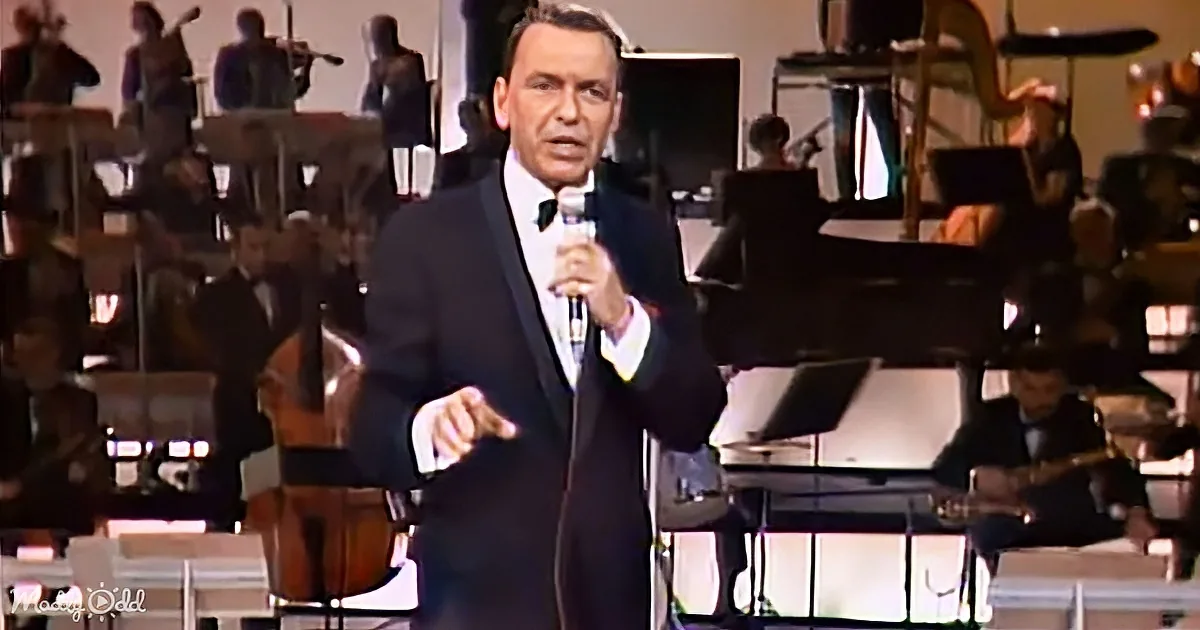Frank Sinatra in a tuxedo sings into a microphone on stage with an orchestra.