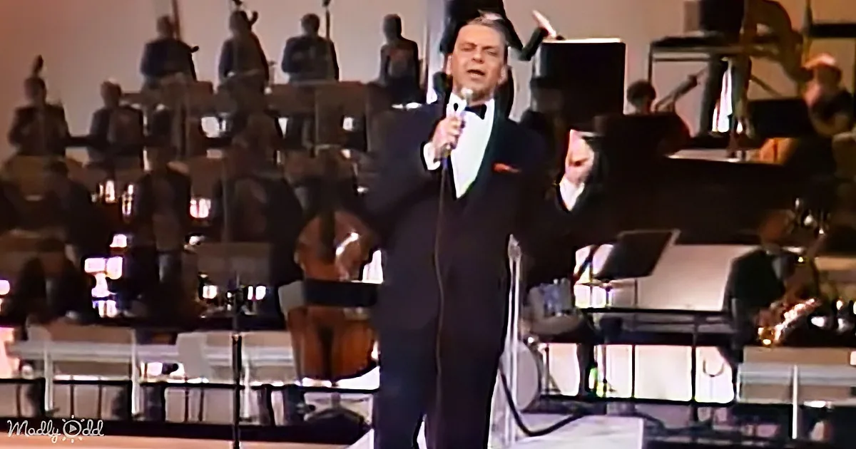 A man in a tuxedo sings into a microphone on stage with an orchestra in the background.