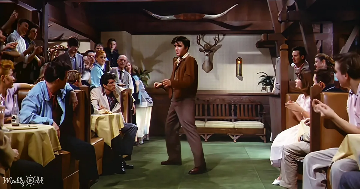 Elvis Presley dances in a restaurant, surrounded by seated patrons and rustic decor.