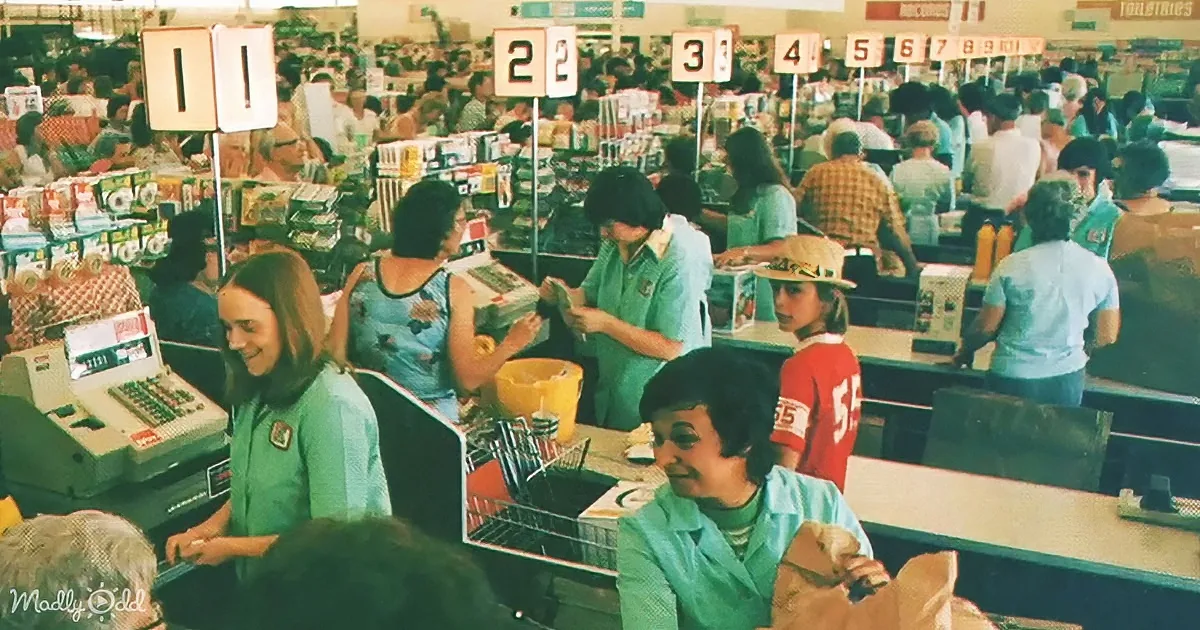 Cashiers in green shirts smile at customers at checkout counters in a bustling grocery store.