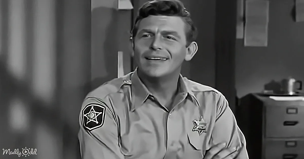 A man in a sheriff's uniform smiles, wearing a badge on his sleeve, in a black-and-white setting.