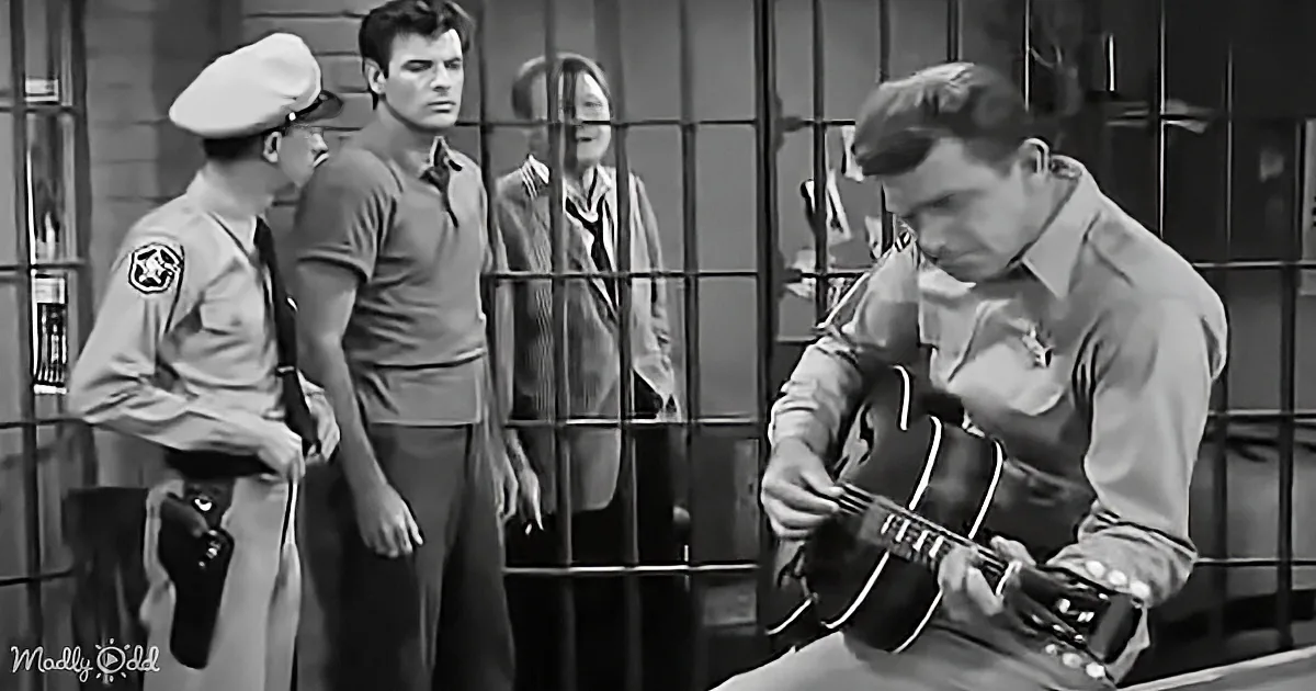 A man in a sheriff's uniform stands beside another man in a jail cell, while a third man plays a guitar.