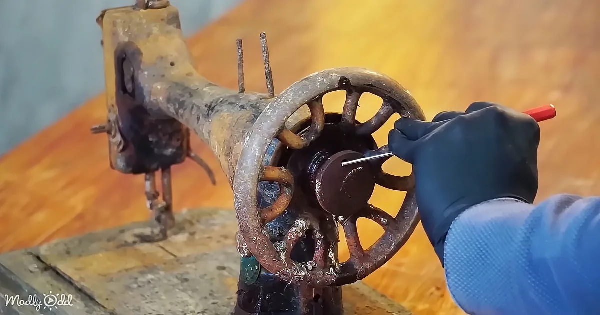 A gloved hand uses a tool to work on a rusty, old sewing machine wheel.