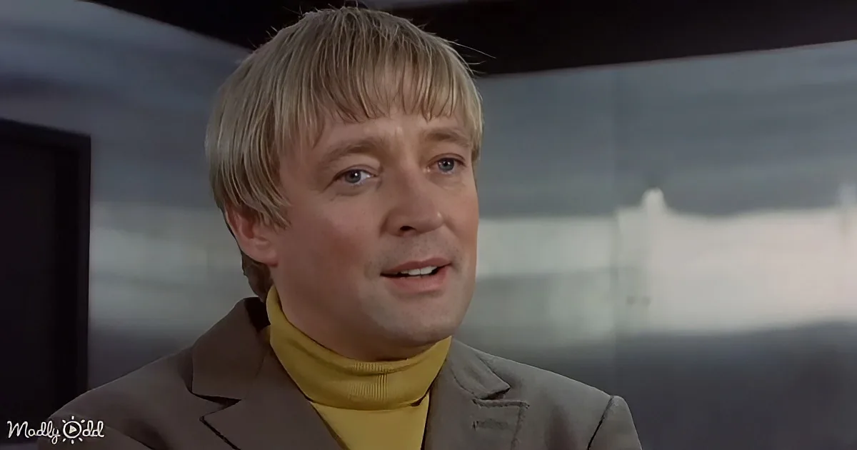 A man with short blonde hair, wearing a yellow turtleneck and a brown blazer, appears to be speaking in a dimly lit room.
